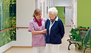 Vanha nainen ja hänestä huolehtiva sairaanhoitaja valaistulla käytävällä